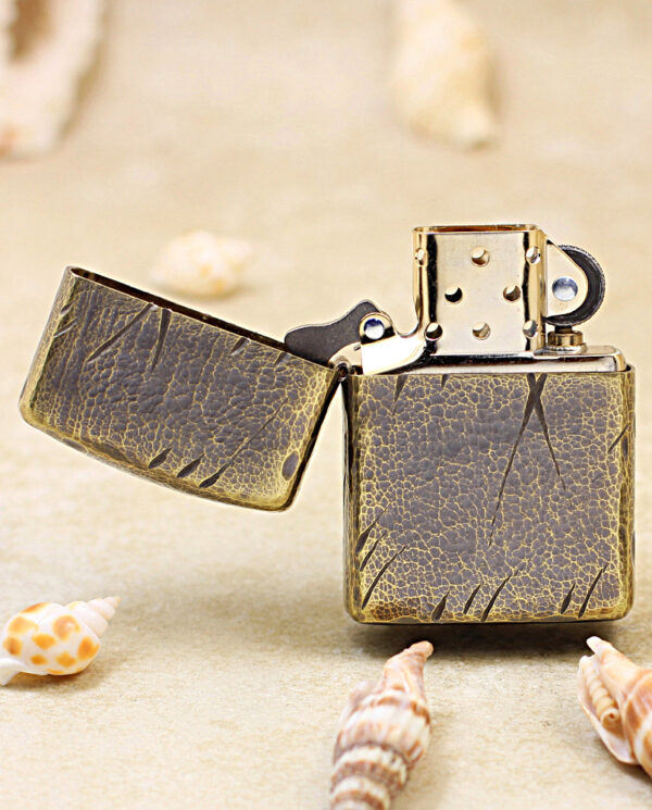 Vintage Zippo-style lighter with cracked patina on sandy surface, surrounded by seashells.
