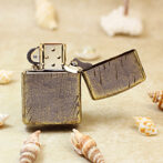Close-up of a vintage leather-wrapped metal lighter with its lid open on a sandy surface, surrounded by seashells.