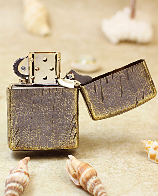 Close-up of a vintage leather-wrapped metal lighter with its lid open on a sandy surface, surrounded by seashells.