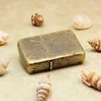 Vintage brass lighter with a cracked patina resting on sandy surface, surrounded by seashells on a beach."],[