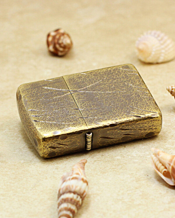Vintage brass lighter with a cracked patina resting on sandy surface, surrounded by seashells on a beach."],[