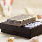 A weathered, cracked Zippo lighter resting on a black jewelry box with seashells around it, on a sandy surface.