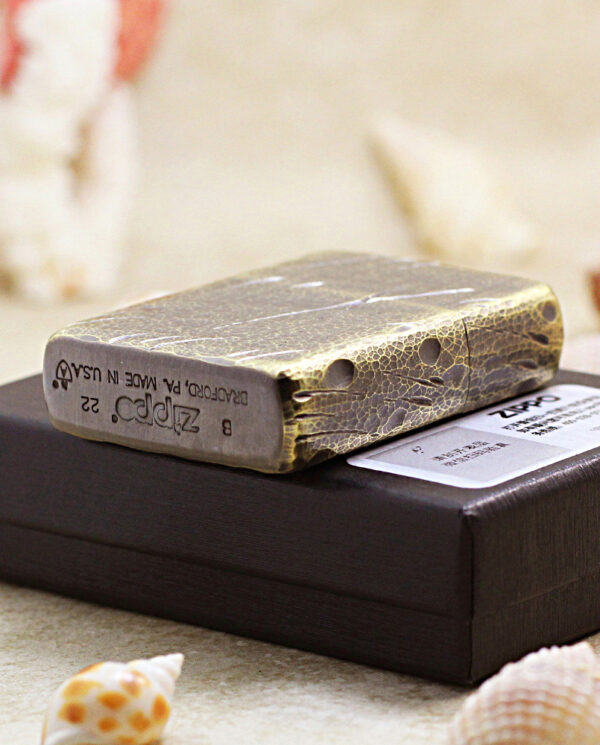 A weathered, cracked Zippo lighter resting on a black jewelry box with seashells around it, on a sandy surface.