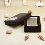 Black presentation box opened to reveal a gold textured rectangular case on a sandy surface with seashells nearby, suggesting a luxury accessory product.