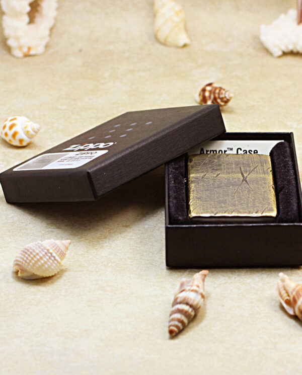 Black presentation box opened to reveal a gold textured rectangular case on a sandy surface with seashells nearby, suggesting a luxury accessory product.