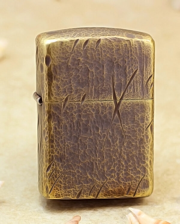 Weathered brass lighter with a cracked patina standing upright on a sandy beach, surrounded by small seashells