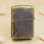 Vintage metal lighter with a distressed bronze finish and engraved scratches, standing upright on a sandy surface.