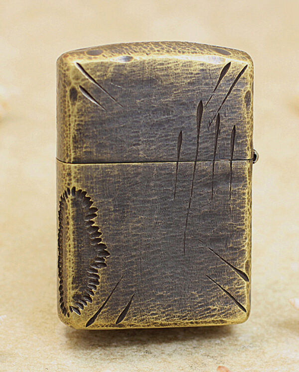 Vintage metal lighter with a distressed bronze finish and engraved scratches, standing upright on a sandy surface.