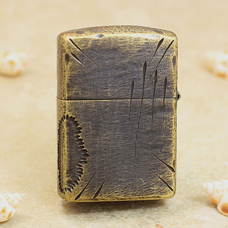 Vintage metal lighter with a distressed bronze finish and engraved scratches, standing upright on a sandy surface.