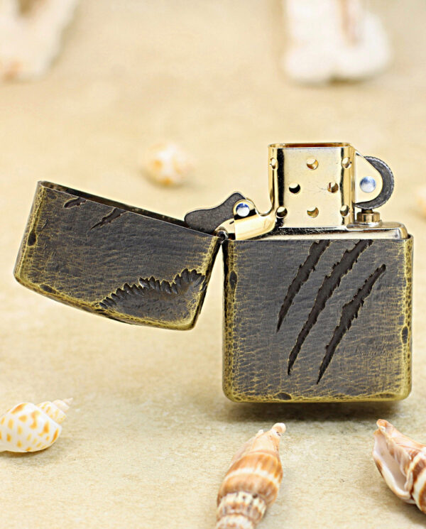 Vintage brass lighter with a dark cracked leather cover and open lid on sandy beach with seashells nearby.