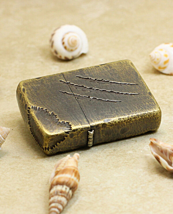 Bronze vintage lighter on sand among seashells, with three scratch marks on its lid.