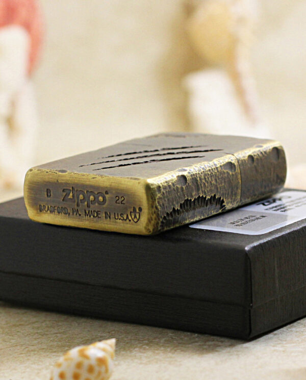 Brass Zippo lighter resting on a black box with seashells in the foreground, close-up shot of the lighter's worn surface.