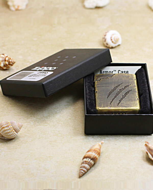 Brass Zippo lighter in a black box labeled Armor Case, surrounded by seashells on a beige surface.