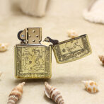 Ornate gold lighter with a hinged lid open, engraved map design on the body, placed on sand with seashells nearby (product photo)