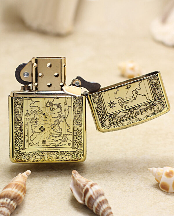 KZ196-03 Ornate gold lighter with a hinged lid open, engraved map design on the body, placed on sand with seashells nearby (product photo)