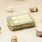Ornate gold-toned lighter with intricate engravings, labeled 'Fire and Blood', resting on a sandy surface among seashells (decorative collectible).