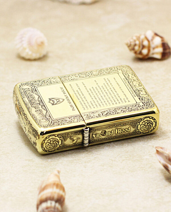 KZ196-04 Ornate gold-toned lighter with intricate engravings, labeled 'Fire and Blood', resting on a sandy surface among seashells (decorative collectible).