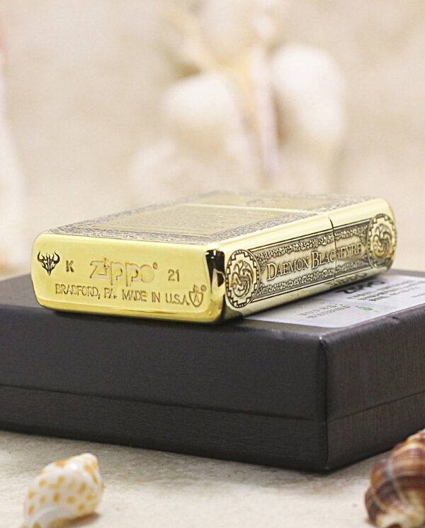 KZ196-05 Gold Zippo lighter with ornate engravings and 'Daemon Black Fire' inscription on a black box, surrounded by seashells.