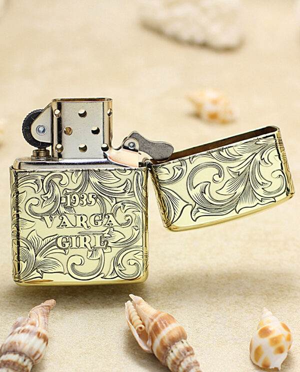 KZ202-03 Ornate metal lighter with '1935 Varga Girl' engraving, open lid, resting on sand among seashells.