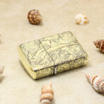 Golden ornate lighter with swirling engravings on a sandy surface, surrounded by seashells.