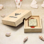 Engraved Zippo lighter in an open wooden presentation box on a sandy surface with seashells nearby.