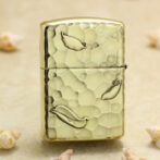 Gold Zippo-style lighter with a hammered textured finish and leaf motifs on a sandy background with seashells nearby.