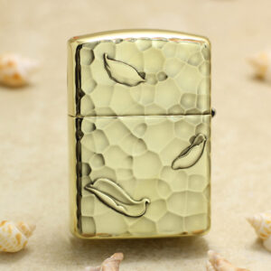 KZ204-01 Gold Zippo-style lighter with a hammered textured finish and leaf motifs on a sandy background with seashells nearby.