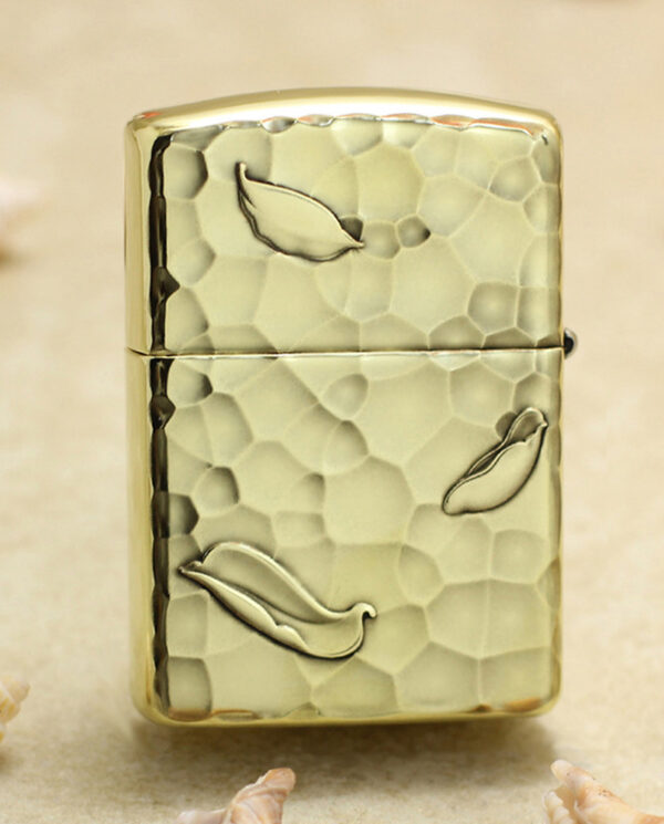 KZ204-01 Gold Zippo-style lighter with a hammered textured finish and leaf motifs on a sandy background with seashells nearby.
