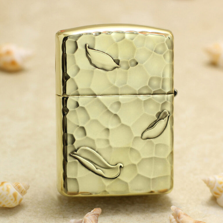 Gold Zippo-style lighter with a hammered textured finish and leaf motifs on a sandy background with seashells nearby.
