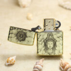 Brass lighter with a hammered pattern and a Buddha relief on the front, open lid, resting on a sandy beach with seashells.