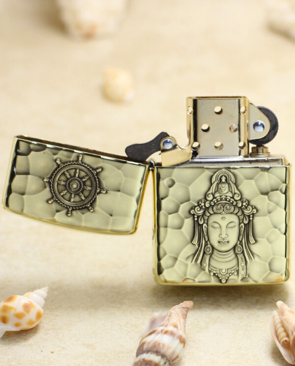 KZ204-02 Brass lighter with a hammered pattern and a Buddha relief on the front, open lid, resting on a sandy beach with seashells.