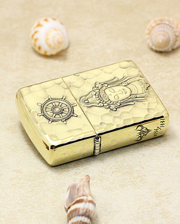 KZ204-04 Gold-tone Zippo-style lighter with embossed ship wheel and ornate figure, placed on sand with seashells nearby