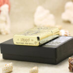 Gold Zippo lighter with engraved design rests on a black box, surrounded by seashells on a sandy surface.