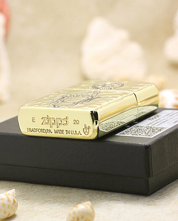 KZ204-05 Gold Zippo lighter with engraved design rests on a black box, surrounded by seashells on a sandy surface.