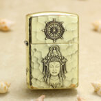 Brass hammered-light metal lighter with a ship-wheel emblem on top and a carved Buddha figure on the body, lying on sand with shells nearby.