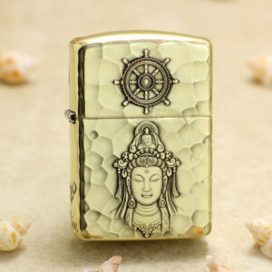 Brass hammered-light metal lighter with a ship-wheel emblem on top and a carved Buddha figure on the body, lying on sand with shells nearby.