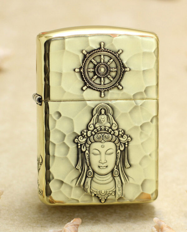 KZ204 Brass hammered-light metal lighter with a ship-wheel emblem on top and a carved Buddha figure on the body, lying on sand with shells nearby.