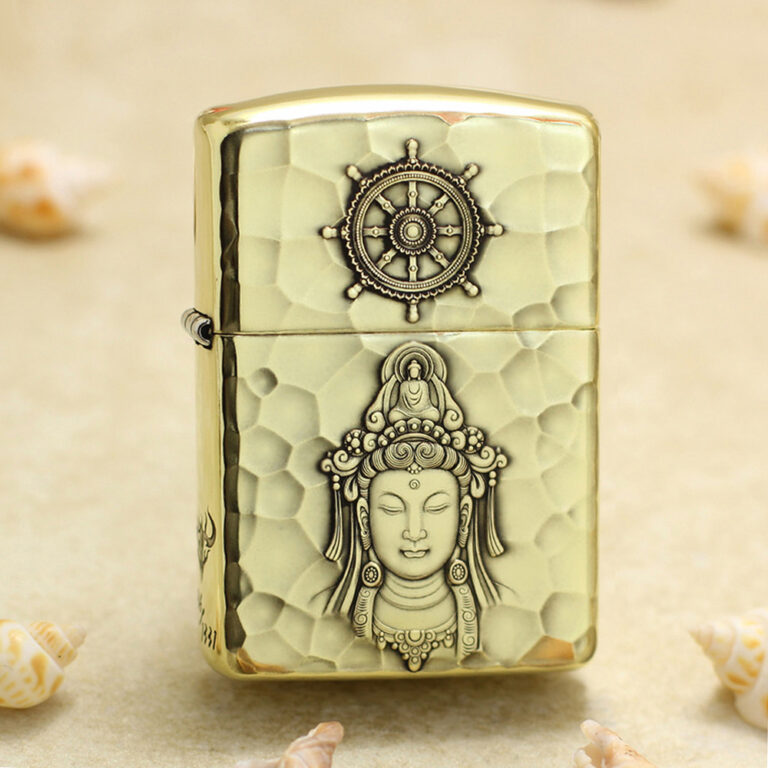 Brass hammered-light metal lighter with a ship-wheel emblem on top and a carved Buddha figure on the body, lying on sand with shells nearby.