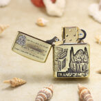 Metal Zippo lighter with Optimus Prime helmet and Transformers logo on a sandy surface with seashells nearby.
