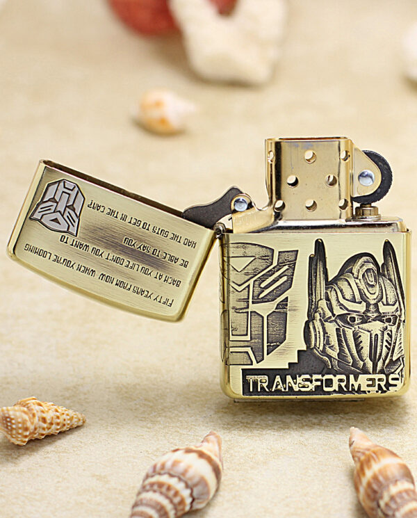 KZ206-02 Metal Zippo lighter with Optimus Prime helmet and Transformers logo on a sandy surface with seashells nearby.