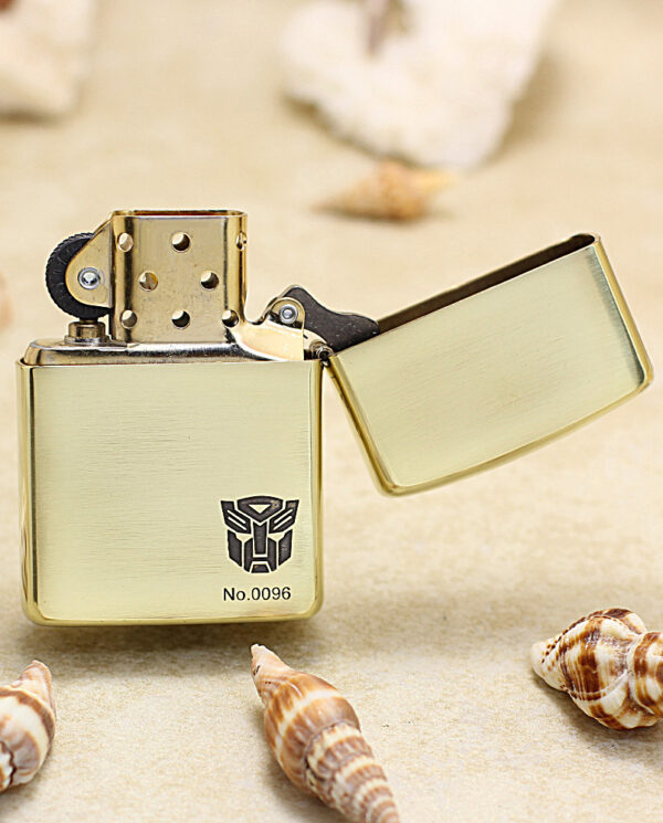 KZ206-03 Gold Zippo lighter with an open lid, featuring an Autobot logo and No.0096 on a sandy background with seashells nearby.
