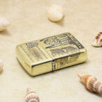 Gold-toned metal lighter with engraved designs rests on sandy beach among seashells.