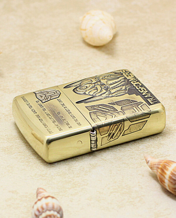 KZ206-04 Gold-toned metal lighter with engraved designs rests on sandy beach among seashells.