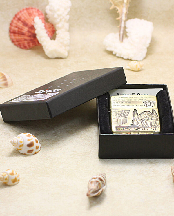 KZ206-06 Transformers Zippo lighter in a black presentation box on a sandy beach with seashells nearby.