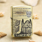 Gold-tone lighter decorated with Transformers artwork, featuring a geometric panel design and a robotic helmet, plus the word Transformers on the front.