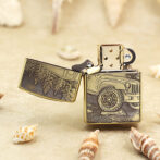 Close-up of a gold Zippo lighter with its lid open, engraved with an off-road jeep design, set on a sandy beach with seashells nearby.