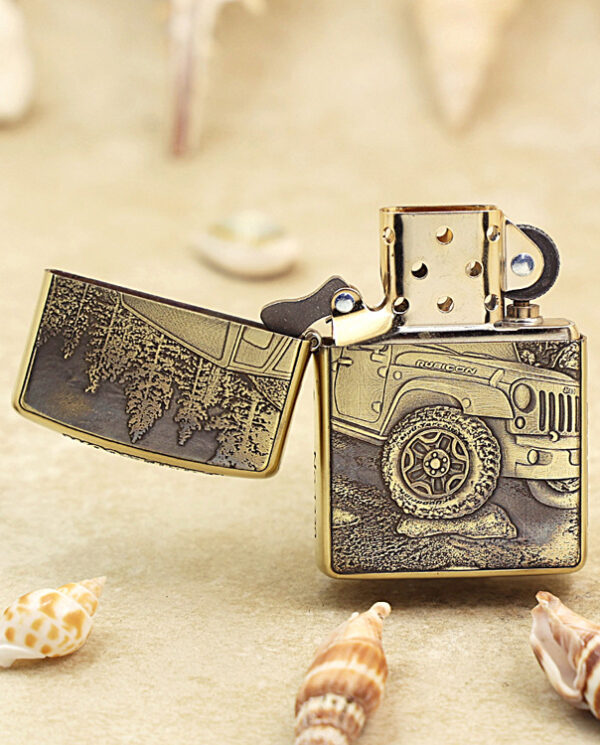 KZ209-02 Close-up of a gold Zippo lighter with its lid open, engraved with an off-road jeep design, set on a sandy beach with seashells nearby.