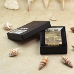 Zippo lighter with a Jeep design inside an open black box on a sandy surface, surrounded by seashells.
