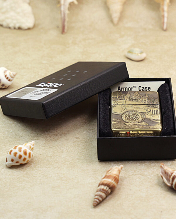 KZ209-06 Zippo lighter with a Jeep design inside an open black box on a sandy surface, surrounded by seashells.