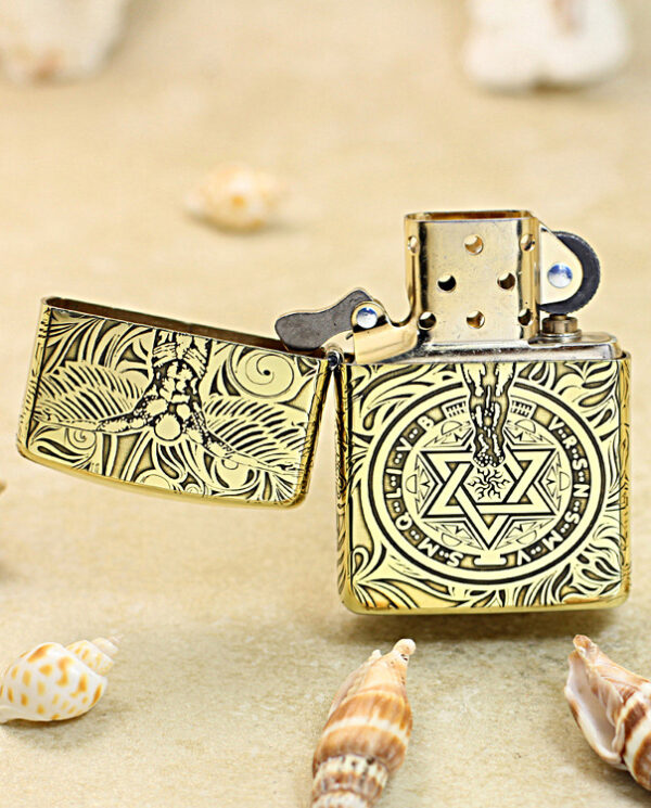 KZ211-02 Decorative brass lighter with intricate swirling patterns, open to reveal its mechanism, on a sandy beach with seashells nearby.